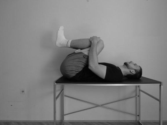 Izvedba: Merjenec leži na hrbtu, z medenico na robu masažne mize. Obe koleni pritegne k prsim, tako da se ledveni del hrbtenice dotika mize.